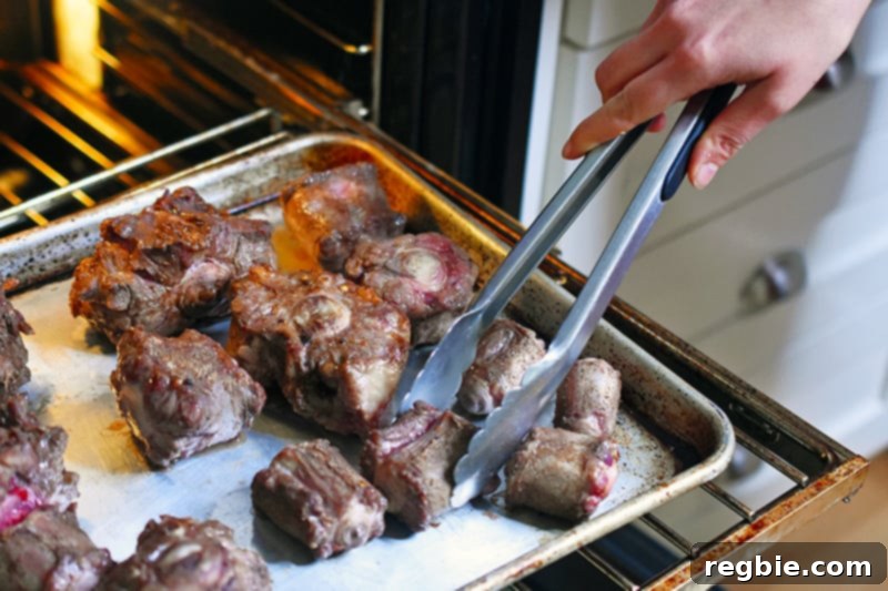 Broiled oxtails being flipped with a pair of tongs to brown evenly.