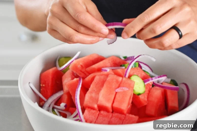Fiesta Watermelon Cucumber Salad 7 Someone adding drained red onions to a large white bowl already containing cubed watermelon and sliced cucumbers.