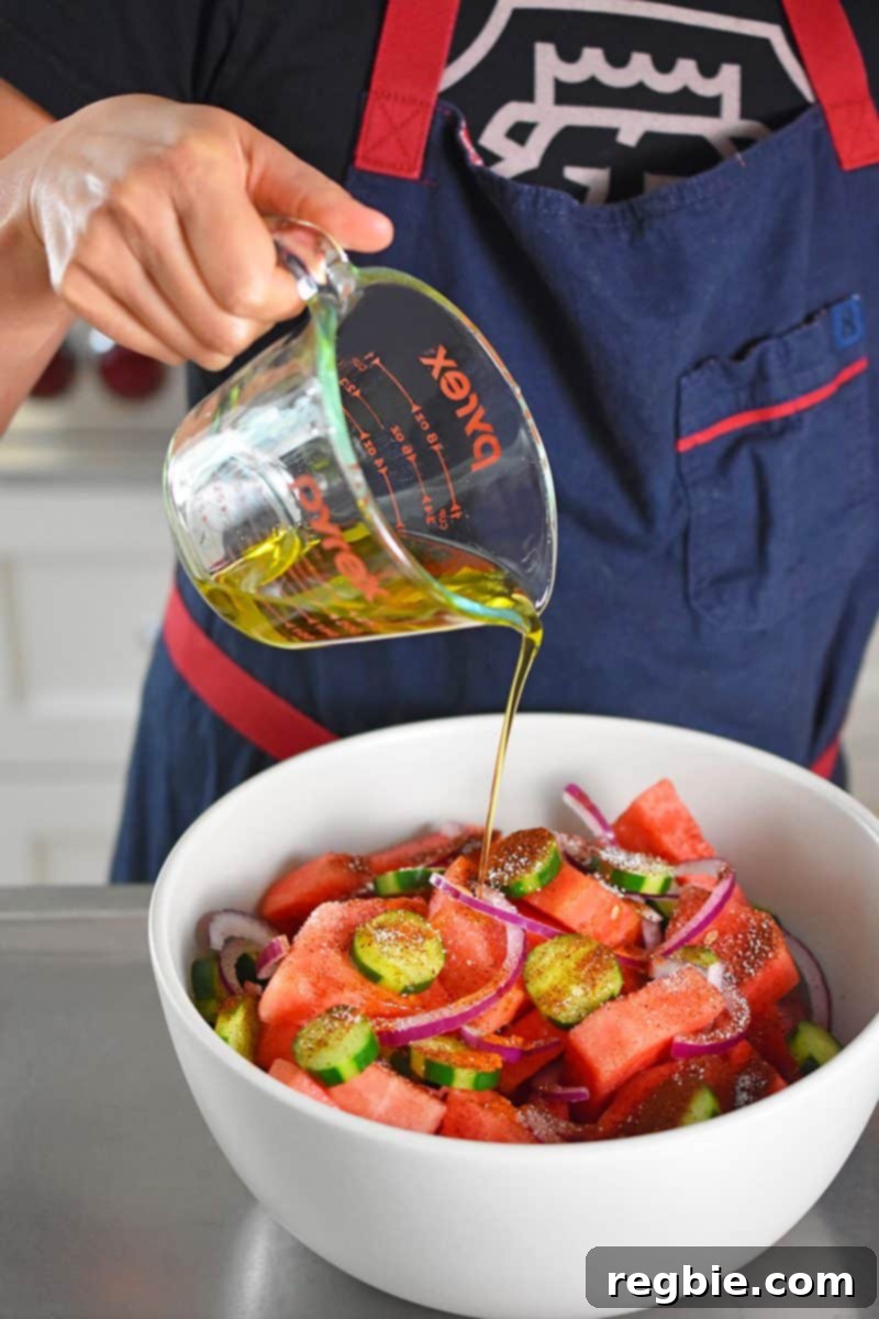 Fiesta Watermelon Cucumber Salad 8 Someone pouring extra virgin olive oil over the watermelon, cucumber, and onion mixture in a white bowl.