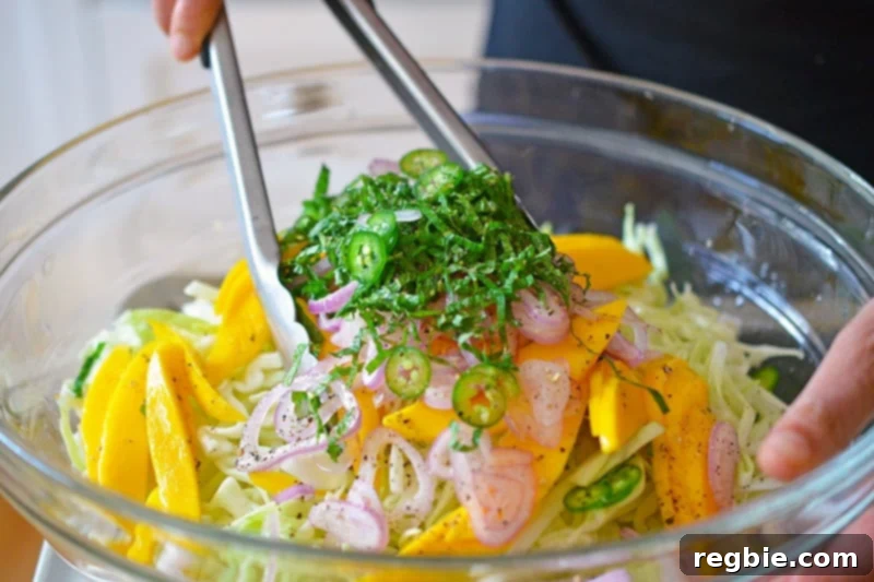 Zesty Mango Cabbage Crunch 8 Someone tossing the spicy mango cabbage slaw with metal tongs in a large glass bowl, ensuring all ingredients are evenly coated.