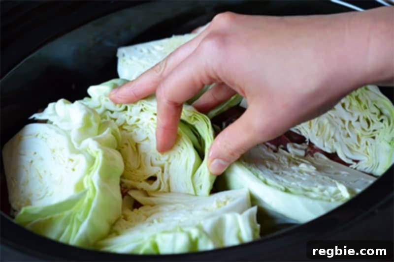 A hand carefully places cabbage wedges on top of the pork and vegetables in an open slow cooker, preparing for the final layer of sauce.