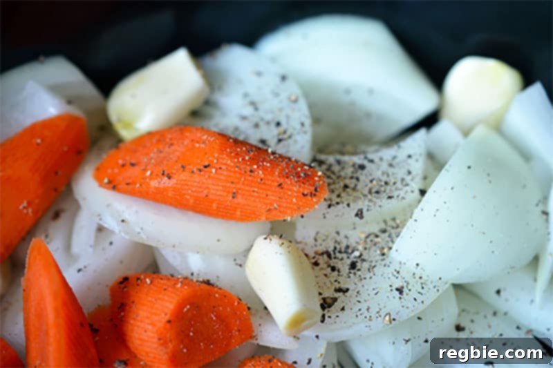 A closeup shot of thinly sliced onions, smashed garlic, and baby carrots seasoned with salt and pepper in the base of a slow cooker, forming the flavorful foundation for the pork stew.