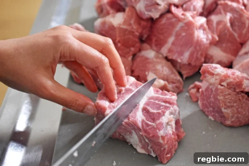 Cutting a pork shoulder roast into uniform 1½-inch cubes on a clean gray cutting board, preparing it for seasoning.