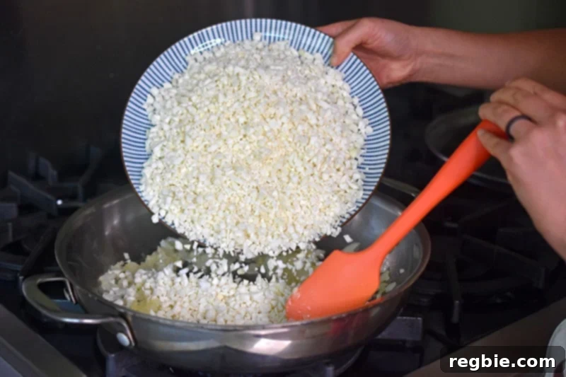 A plate of freshly riced cauliflower is being poured into the skillet, ready to be transformed into Whole30 Mexican Cauliflower rice.