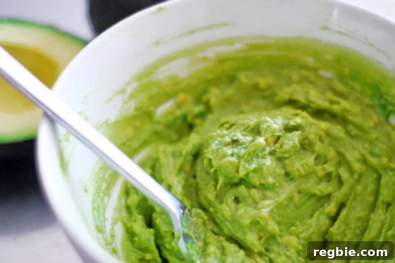 Avocado Dream Dip 5 A white bowl containing half of the avocado mashed with a fork, exhibiting a creamy texture and vibrant green color.