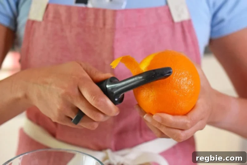 A person in a red apron is peeling the zest off of an orange with a vegetable peeler.