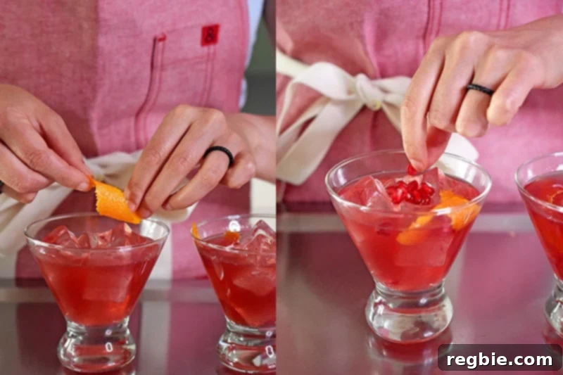 A person in a red apron is adding a piece of twisted orange zest and pomegranate arils to a glass filled with a pomegranate orange juice mocktail.