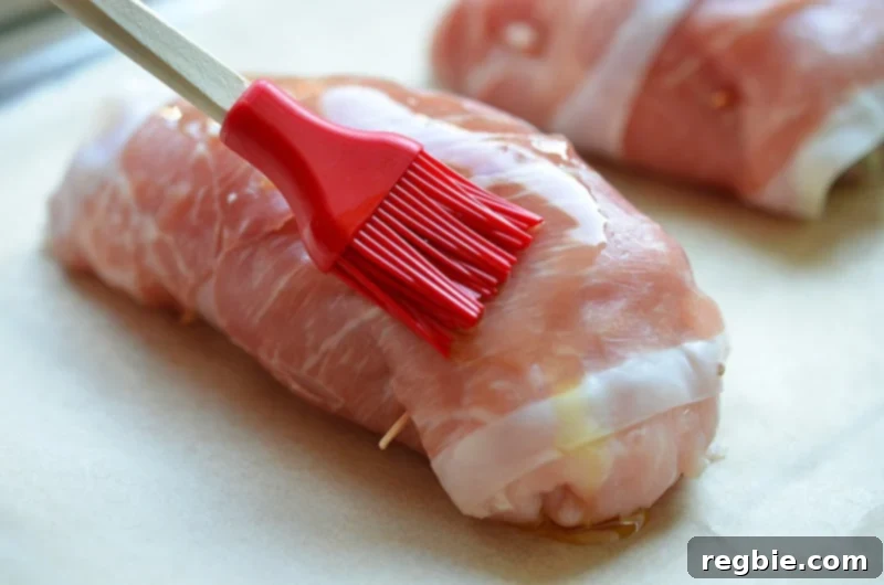 Delectable Prosciutto Chicken Rolls 11 Extra virgin olive oil is being brushed delicately over the tops of the Chicken Prosciutto Involtini with a vibrant red silicone brush. This crucial step enhances the crispness of the prosciutto, contributes to a beautiful golden-brown finish, and ensures an even, appetizing bake during cooking.