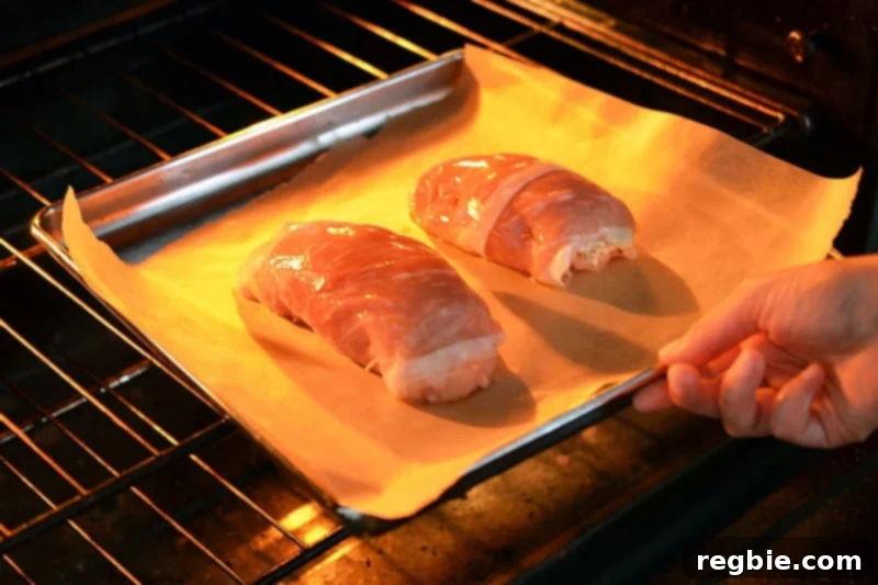 Delectable Prosciutto Chicken Rolls 12 Two neatly prepared Chicken Prosciutto Involtini, glistening slightly with olive oil, are carefully placed on a parchment-lined baking sheet and inserted into a preheated oven. The image captures the anticipation of a delicious meal, showing the dish poised for its transformation into golden perfection.