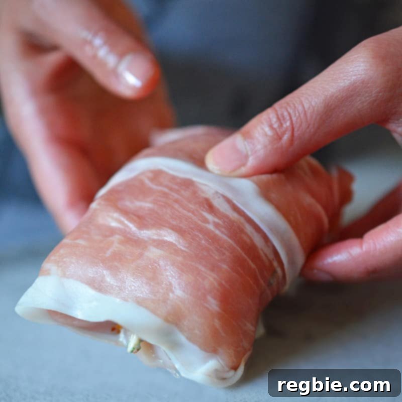 Delectable Prosciutto Chicken Rolls 3 A person is meticulously wrapping a flattened chicken breast with two slices of prosciutto, preparing it for baking. The image highlights the careful preparation and artisanal quality of the dish, emphasizing the hands-on process of creating this impressive and flavorful meal.