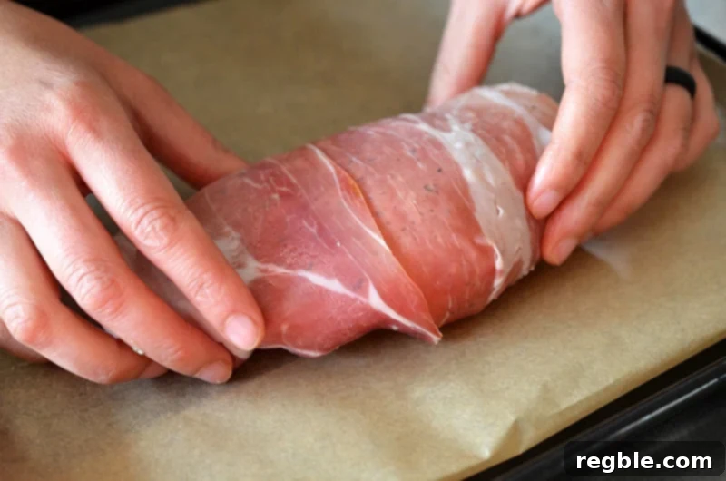 Delectable Prosciutto Chicken Rolls 10 Two slices of delicate prosciutto are expertly wrapped around each rolled chicken packet, securely encasing the savory filling within. The involtini are then carefully placed onto a parchment-lined baking sheet, meticulously arranged and ready for the final touch before baking: a light brush of extra virgin olive oil.