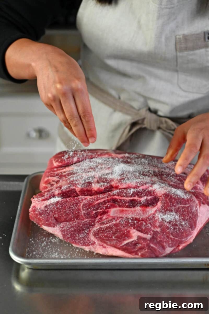 Effortless Peach Glazed Pork Roast 5 A hand is sprinkling kosher salt all over a large bone-in pork shoulder roast.