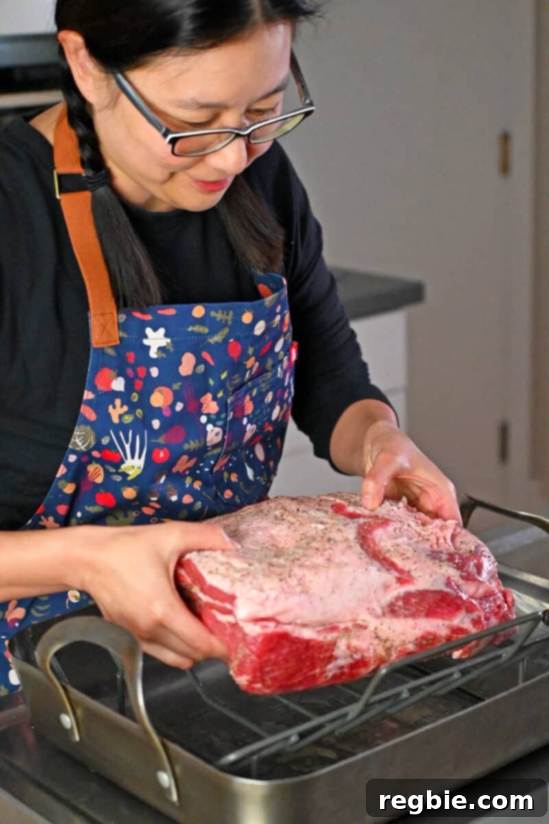 Effortless Peach Glazed Pork Roast 8 An Asian woman is placing a seasoned bone-in pork shoulder roast onto a greased rack in a roasting pan.