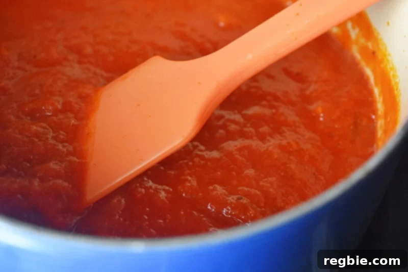 A close-up of a silicone spatula stirring vibrant red marinara sauce in a small Le Creuset saucepan on a stovetop, warming it gently.