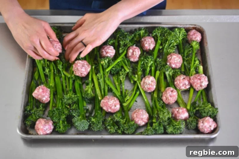 Two hands are shown adding newly formed meatballs to a rimmed baking sheet, placing them strategically among the broccolini.
