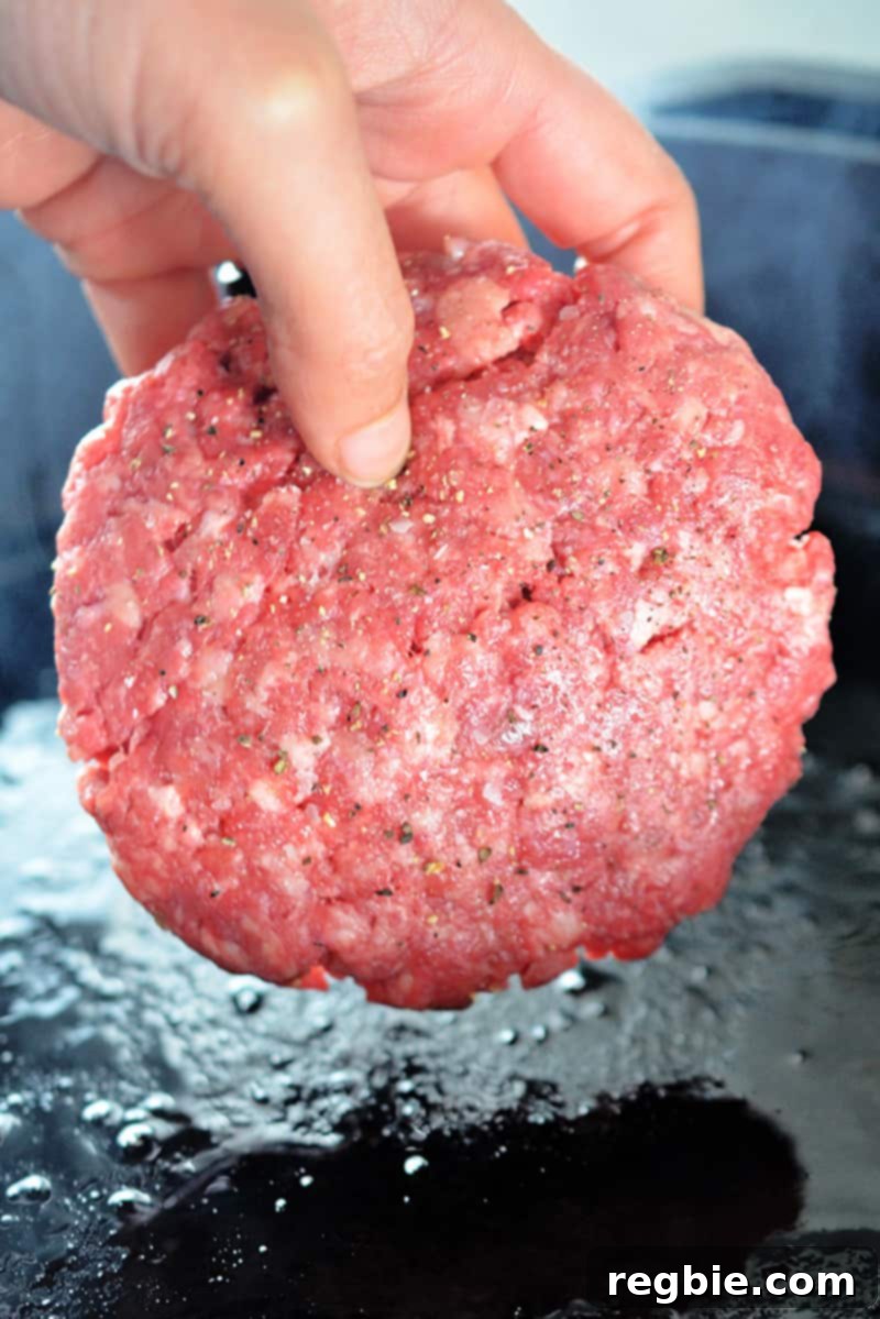 A hand is carefully placing a seasoned Nomster Burger into a sizzling hot cast iron skillet.