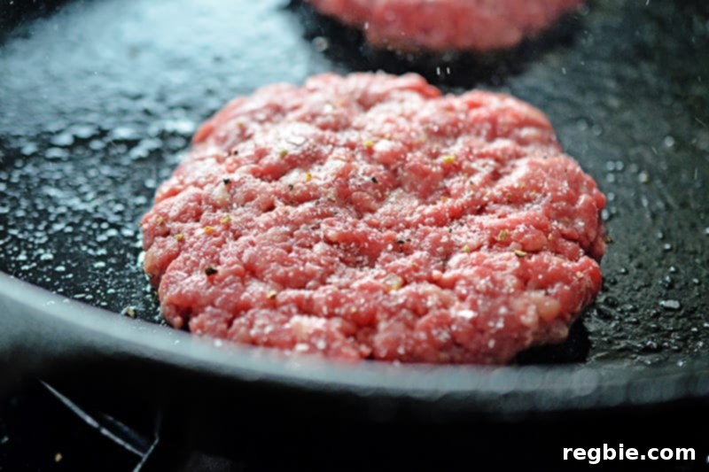 A closeup of Nomster burgers sizzling and frying in a cast iron skillet, with salt and pepper visible on the top.