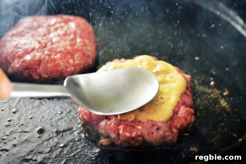 A spoon is spreading Dijon-style mustard evenly on top of a Nomster burger patty frying in a cast iron skillet.