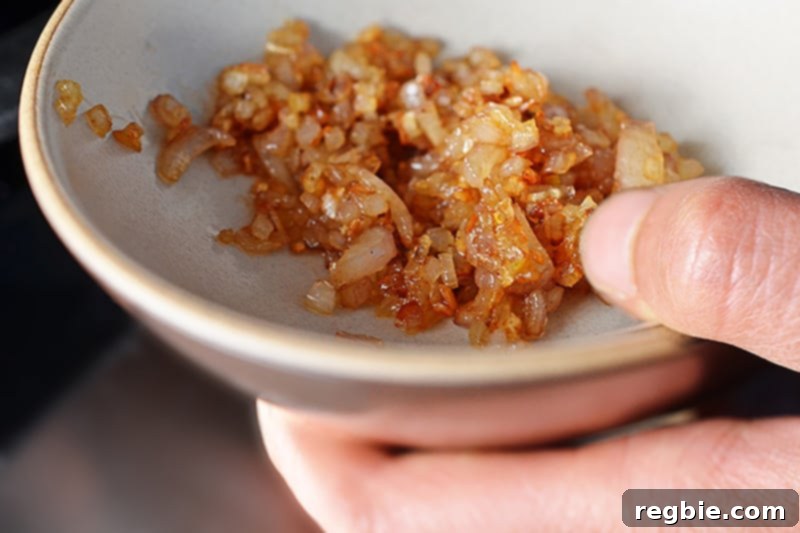 A hand holding a shallow bowl filled with perfectly caramelized diced shallots, cooled and ready for the next step.