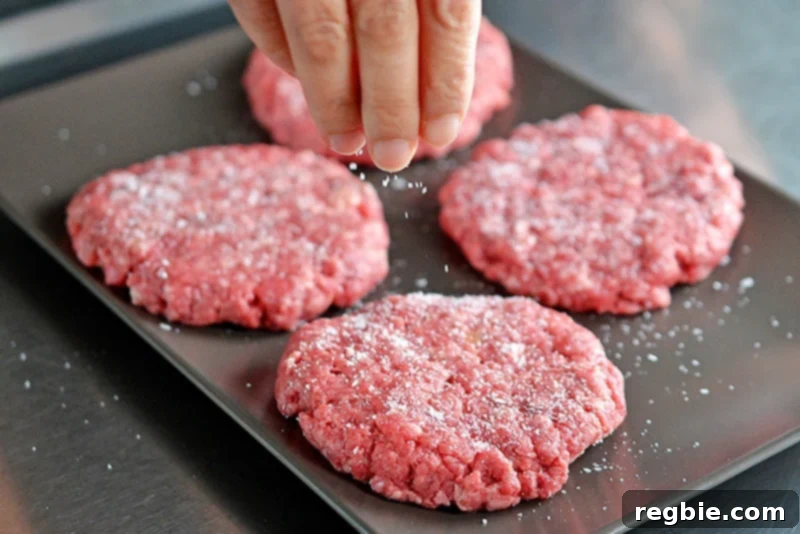 A hand is sprinkling kosher salt generously on four Nomster Burgers, preparing them for the skillet.
