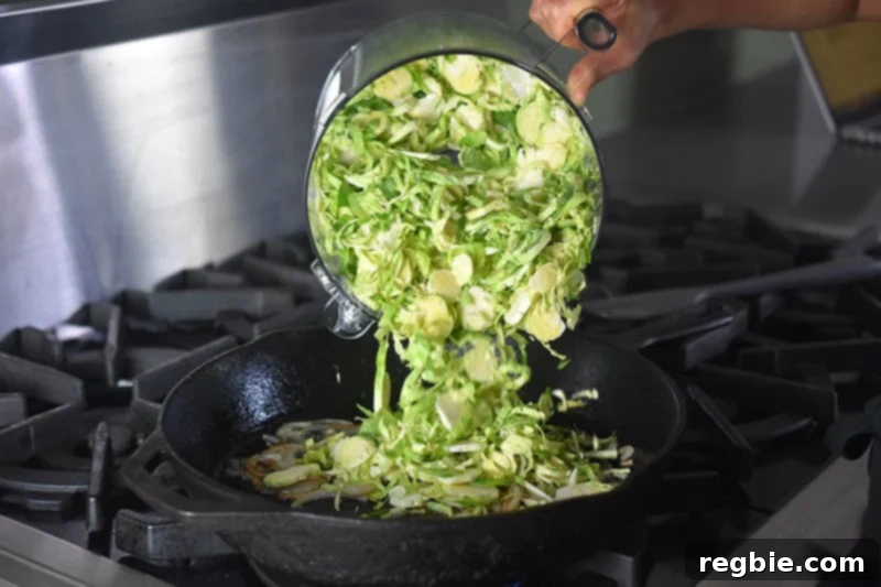 Crispy Bacon Brussels Sprouts 9 Freshly shaved Brussels sprouts being added from a food processor container into a large cast iron skillet with sautéed shallots.