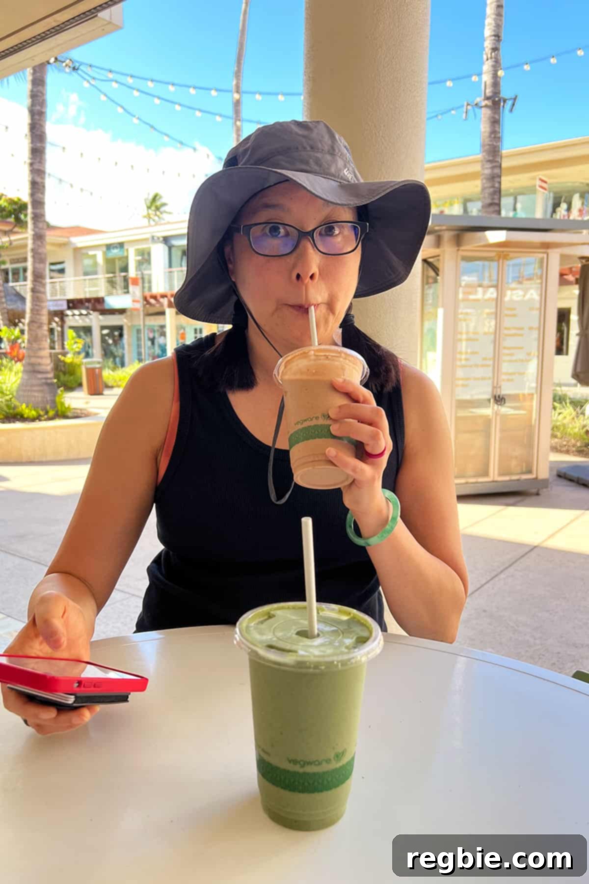 An Asian woman in a hat is drinking a brown smoothie while holding a red cell phone.