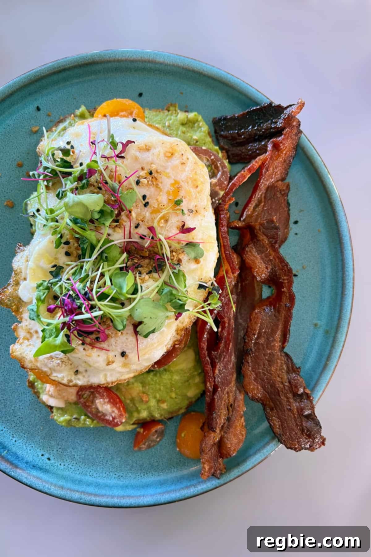 A gluten free avocado toast topped with fried eggs with bacon on the side.