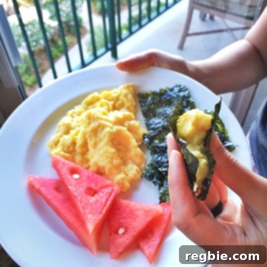A hand is holding a white plate filled with watermelon slices, scrambled eggs, and toasted seaweed.