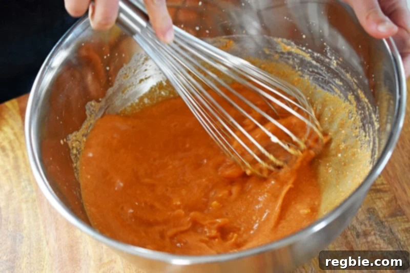 A hand whisking the vibrant orange curry sauce vigorously in a large metal mixing bowl, ensuring all ingredients are fully incorporated.