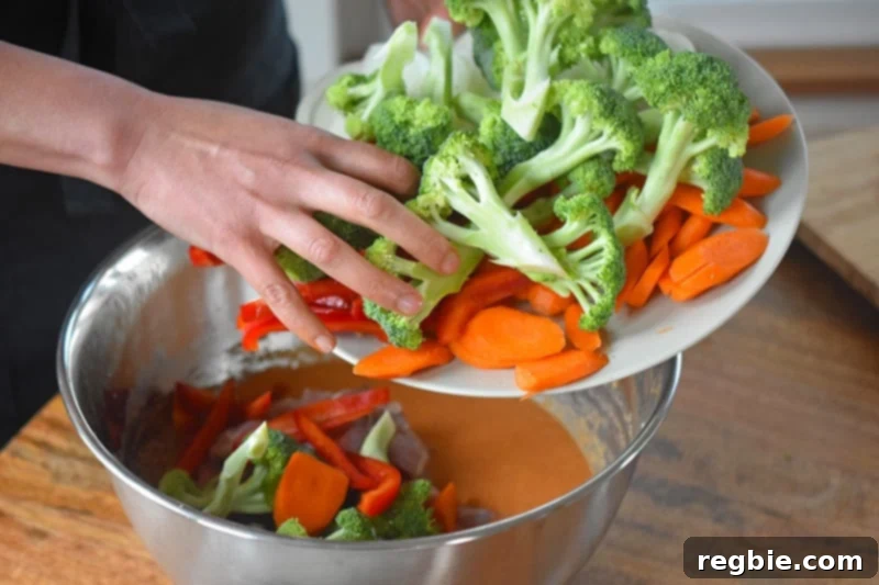 Cut-up broccoli florets, carrot slices, and red bell pepper strips mixed with sliced chicken in the curry sauce within a large metal bowl.