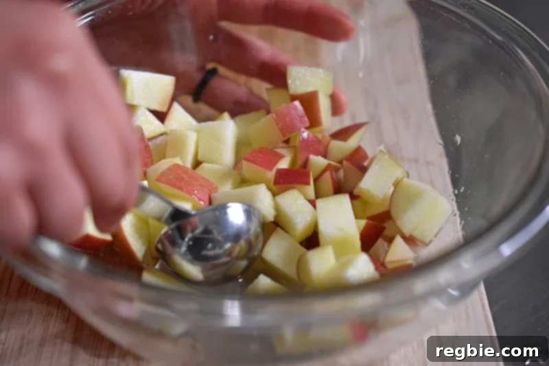 A small spoon is used to gently toss chopped apple cubes with lemon juice in a bowl, ensuring even coating.