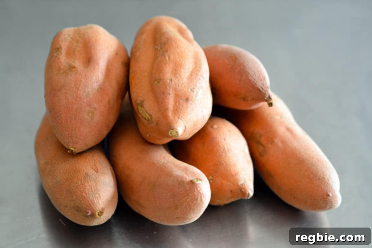 A pile of fresh garnet yams or red-fleshed sweet potatoes resting on a clean stainless steel counter, ready for preparation.