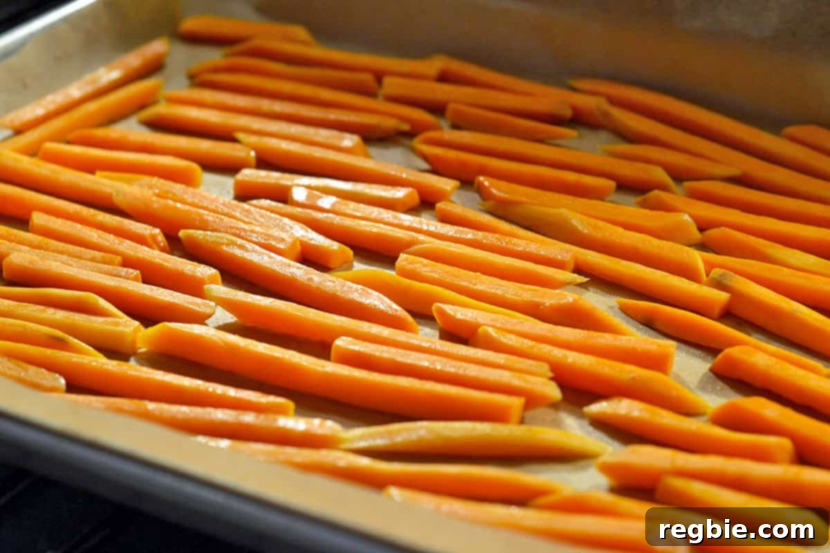 Sweet potato fries, seasoned and coated in oil, are spread in a single layer on a parchment-lined rimmed baking sheet, ready for the oven.