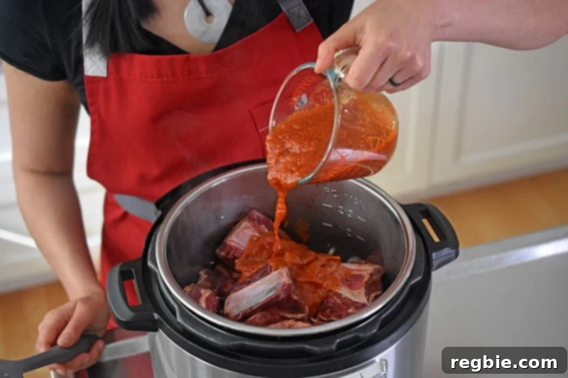 Pouring marinara from a liquid measuring cup over the seasoned short ribs in the Instant Pot, ensuring they are well-coated.