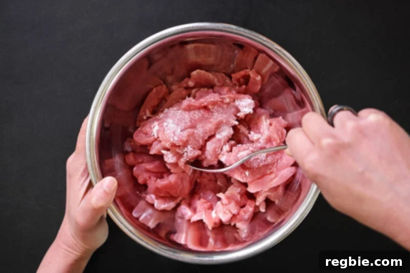 An overhead shot of two hands mixing sliced pork tenderloin strips with salt and avocado oil in a silver bowl.