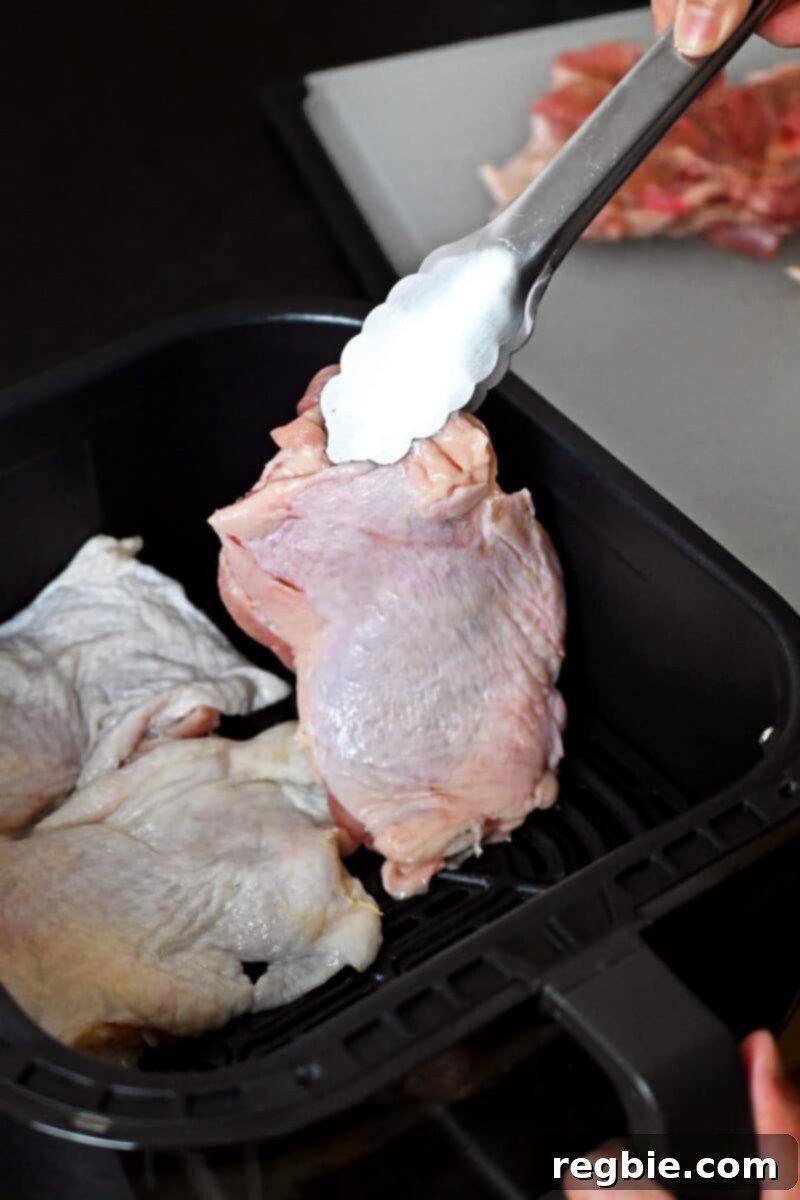 Ultimate Crispy Air Fryer Chicken Thighs 8 A pair of tongs carefully places a raw, seasoned chicken thigh into an air fryer basket, skin-side up.