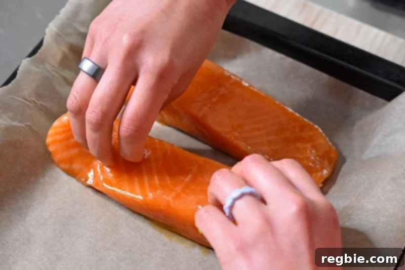Two hands carefully placing marinated teriyaki salmon fillets, skin-side down, on a parchment paper-lined baking sheet.