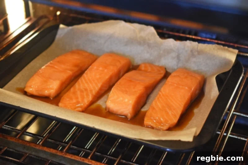 Four teriyaki salmon fillets are perfectly baking on a sheet pan in the oven, creating a delicious aroma.