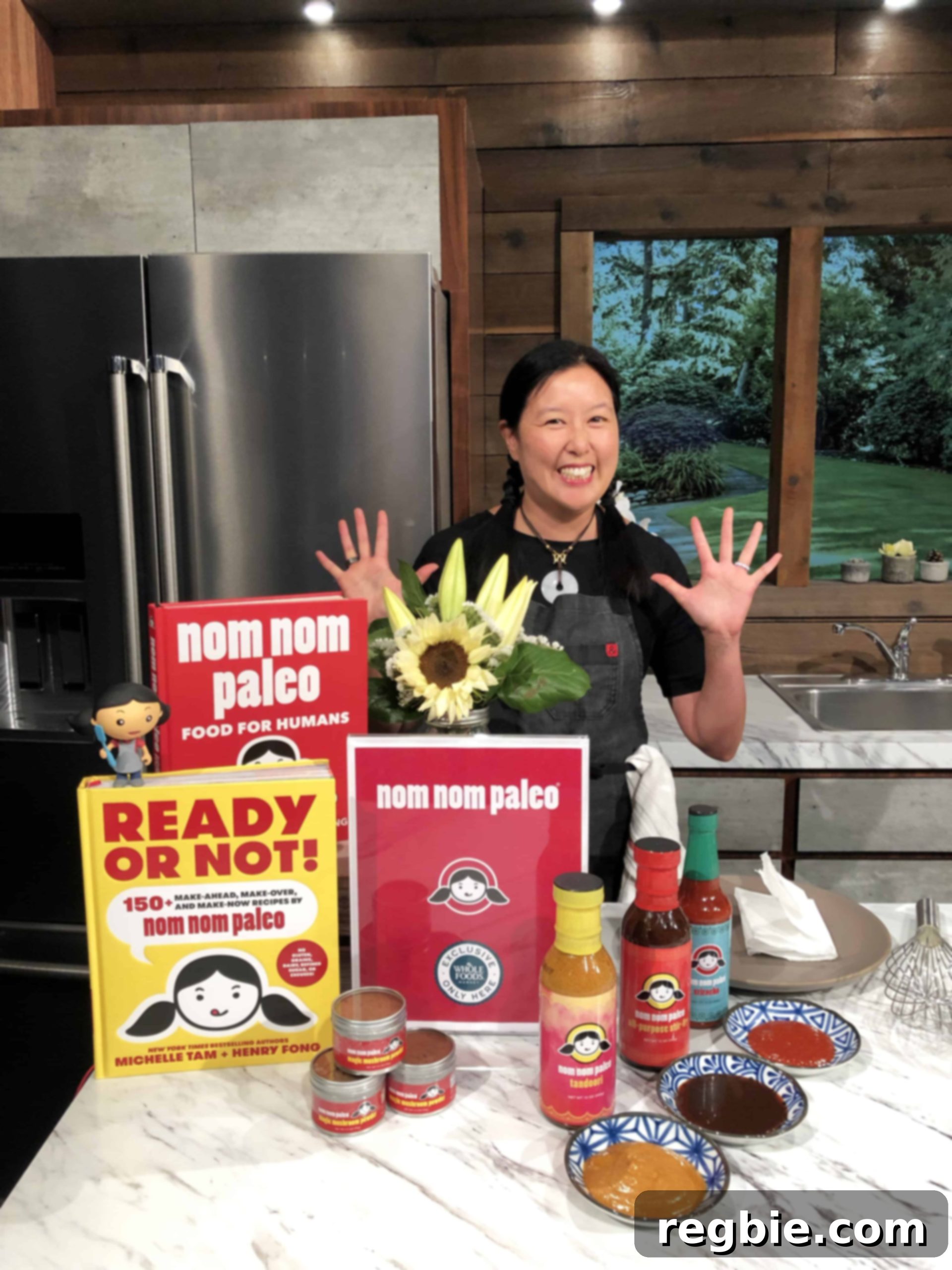 Nom Nom Paleo's Irresistible Sauces 7 Michelle Tam standing in a kitchen behind a display of her Nom Nom Paleo cookbooks, sauces, and Magic Mushroom Powder.