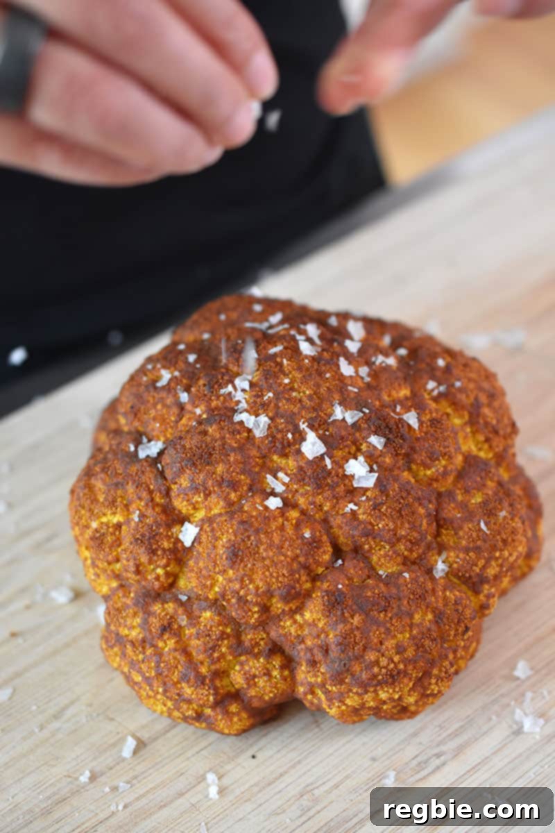 An overhead shot of someone delicately sprinkling flake finishing salt over the golden-brown Tandoori Whole Roasted Cauliflower, enhancing its presentation and flavor.
