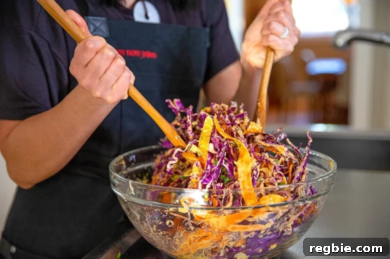 Bright Red Cabbage Slaw with Zesty Ginger Carrot Dressing 7 Someone skillfully tossing Whole30-friendly red cabbage slaw in a large clear mixing bowl using two wooden spoons, ensuring the dressing evenly coats all ingredients.