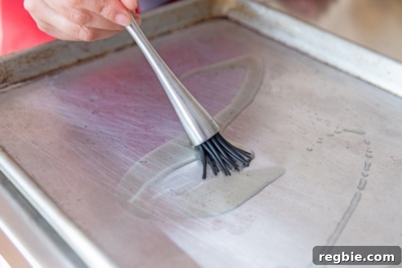 A silicone brush is spreading avocado oil on a rimmed baking sheet for a healthy sheet pan chicken dinner.