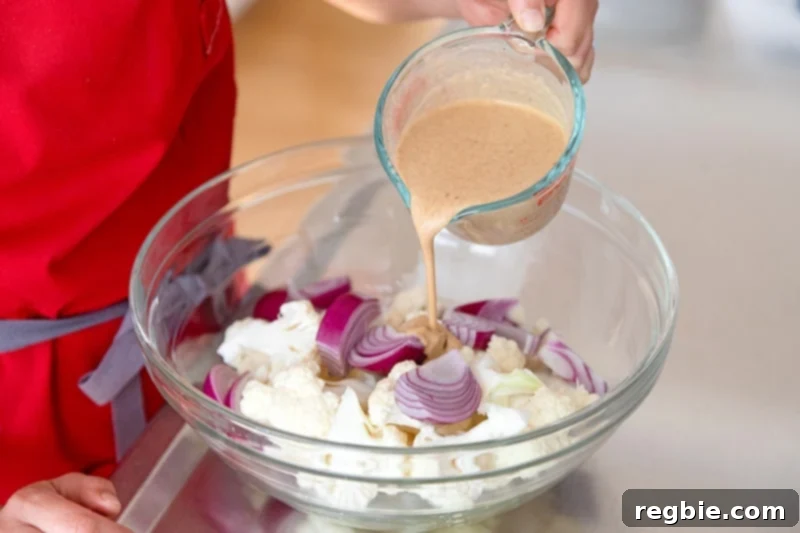 Pouring dairy-free tandoori marinade on cauliflower and onions in a large bowl for Sheet Pan Tandoori Chicken