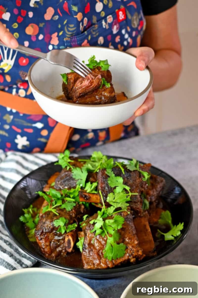 Fall-Off-The-Bone Slow Cooker Korean Short Ribs 3 A person in a biue apron is holding a bowl of braised Korean Short Ribs above a serving bowl of the ribs.