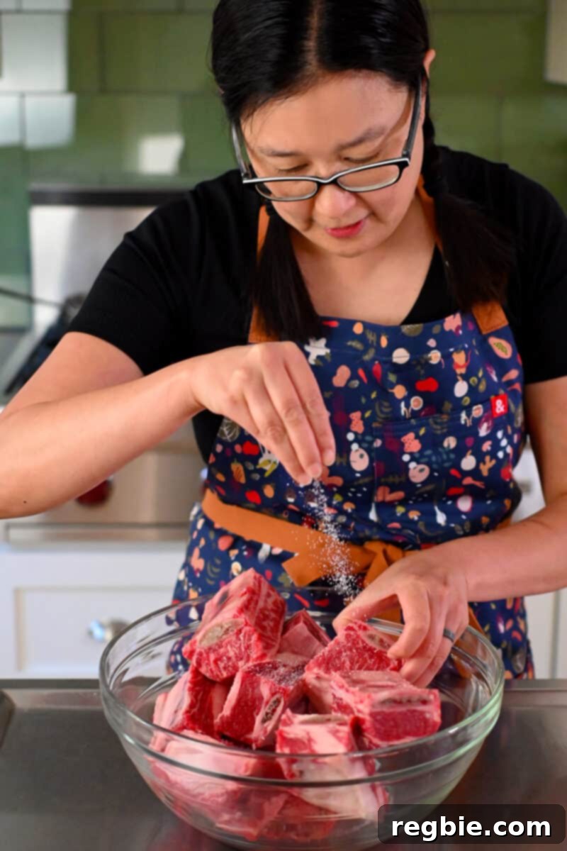 Fall-Off-The-Bone Slow Cooker Korean Short Ribs 7 An Asian woman in glasses is seasoning raw beef short ribs with salt and pepper.