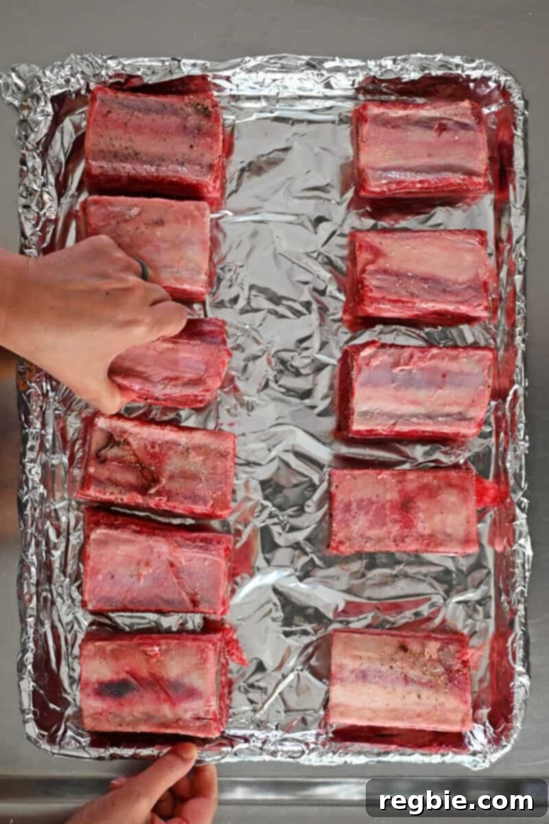 Fall-Off-The-Bone Slow Cooker Korean Short Ribs 8 A hand is placing seasoned raw beef short ribs, bone-side up, on a foil lined rimmed baking sheet.