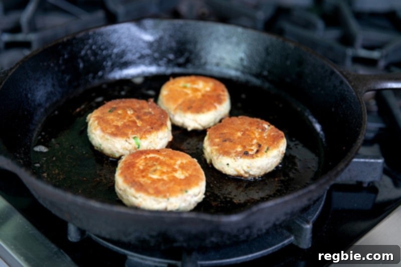 Four golden brown gluten-free crab cakes frying in a cast iron skillet.