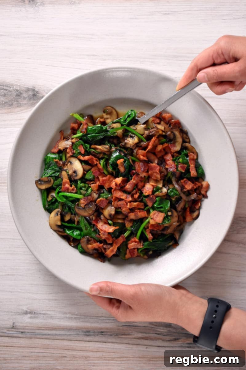 Sizzling Bacon, Mushroom, and Spinach Skillet 15 Two hands are holding a cream colored bowl filled with sautéed spinach with bacon and mushrooms.