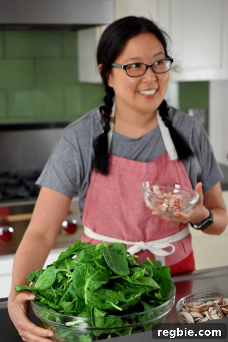 Sizzling Bacon, Mushroom, and Spinach Skillet 4 An Asian woman, wearing a vibrant red apron, smiles as she holds a large bowl brimming with fresh, raw spinach and a separate small bowl of thinly sliced raw bacon, ready to begin preparing a delicious meal.