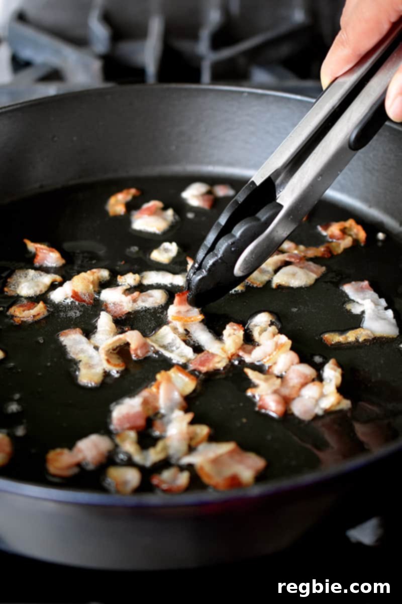 Sizzling Bacon, Mushroom, and Spinach Skillet 6 A pair of tongs gently pushes raw bacon bits around in a large cast iron skillet, while a metal slotted spoon transfers crispy bacon bits onto a paper towel-lined plate.